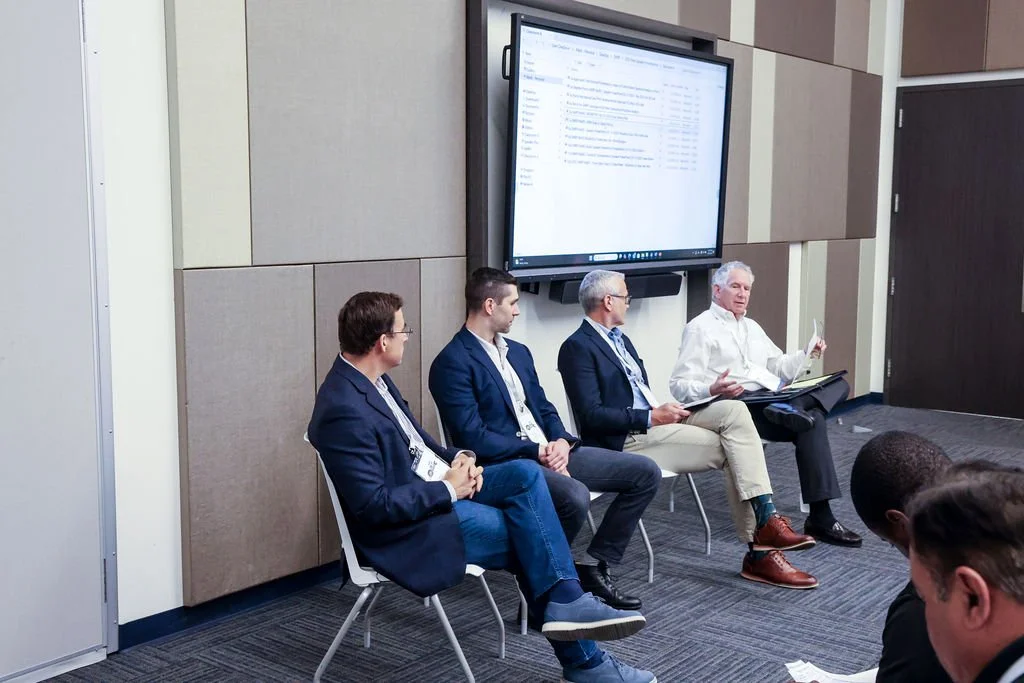 A panel of five men seated on chairs in a conference room, with one man speaking. A large screen behind them displays a document or email list.