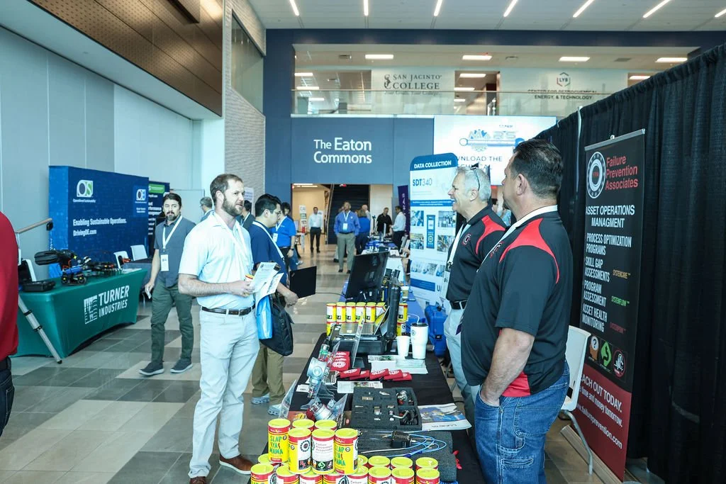 Attendees at a trade show booth engaging in conversation, with displays of products and informational posters in a modern convention center.