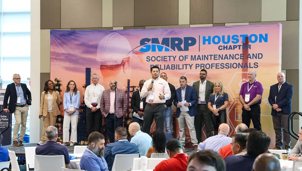 A group of professionals on stage at a conference, with a large banner behind them reading 'SMRP Houston Chapter Society of Maintenance and Reliability Professionals.' An audience is seated in front of the stage.