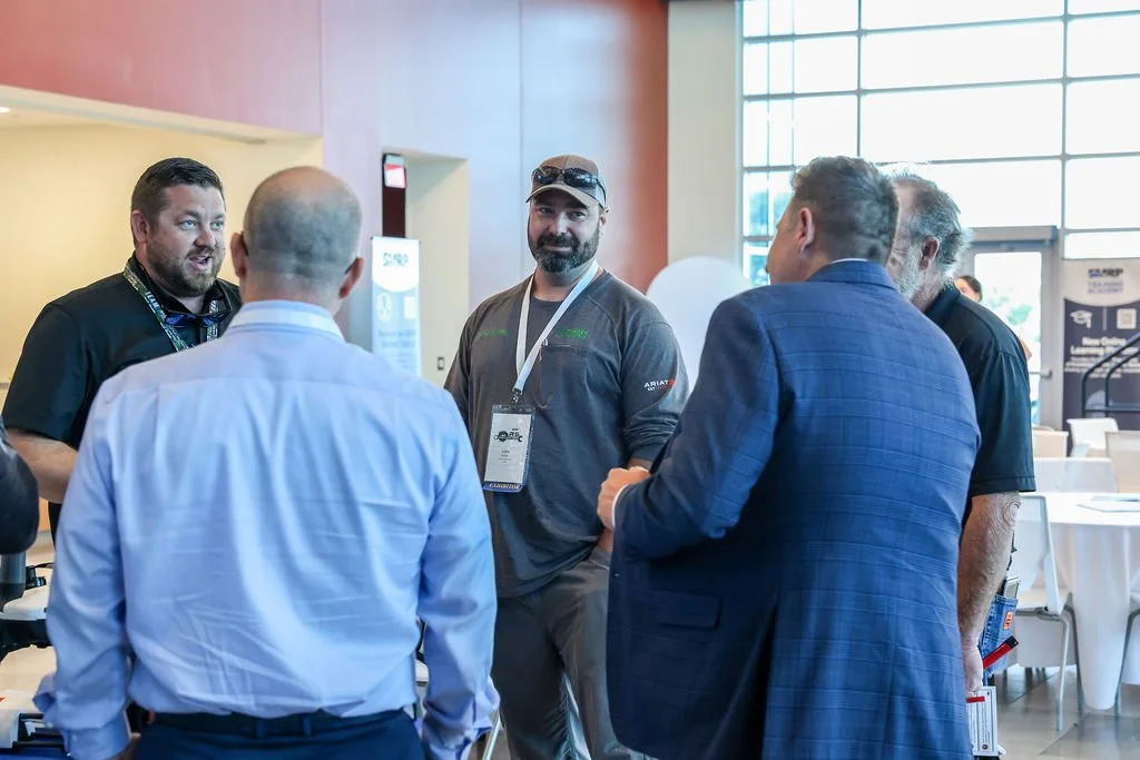 A group of five men having a conversation at a conference, with big windows in the background letting in natural light.