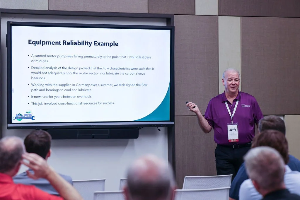 A man giving a presentation in front of a screen with a slide titled 'Equipment Reliability Example' at a conference.