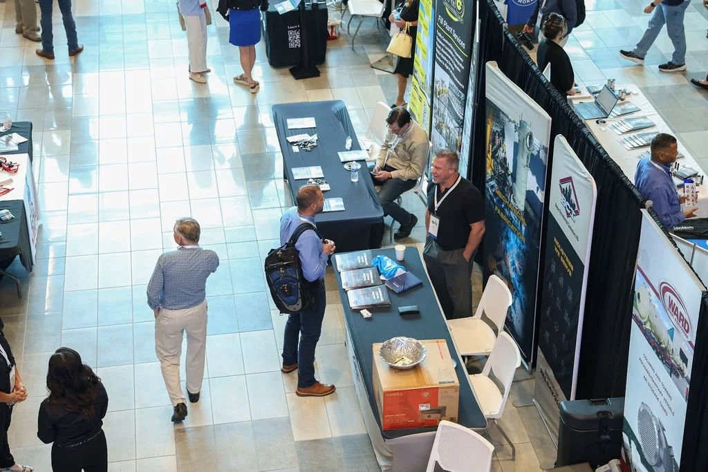 People at a trade show or convention in an indoor mall or exhibition center, engaging in conversations near display booths with banners and promotional materials.