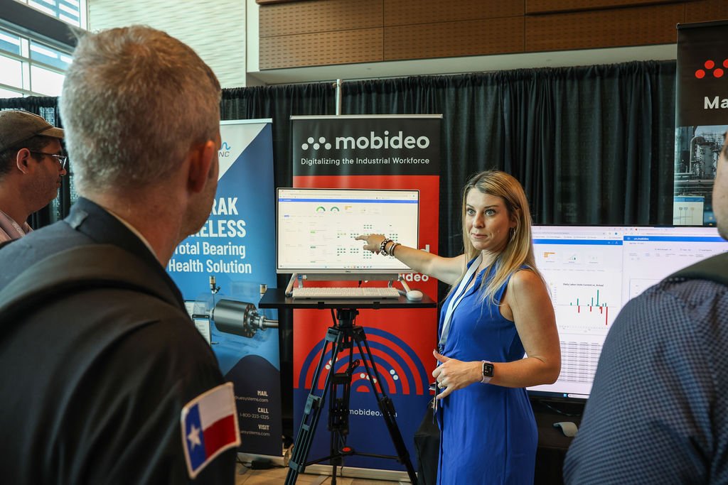 Woman in blue dress explaining data on a monitor at a trade show booth for Mobideo, with two men listening nearby.