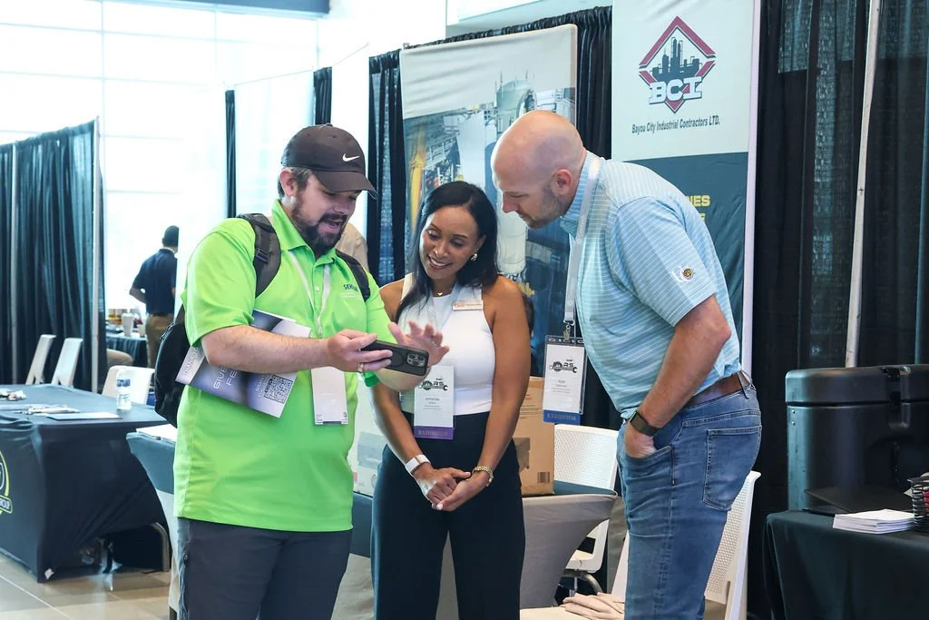Three people are discussing and looking at a phone at a trade show or conference. Behind them are display banners, tables, and chairs.