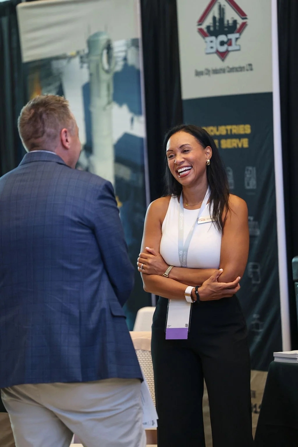 Two people talking at a conference booth, a woman smiling and a man with his back turned. The woman is wearing a white sleeveless top and black pants, with lanyards. The booth banners behind them display logos and text about industries and services.