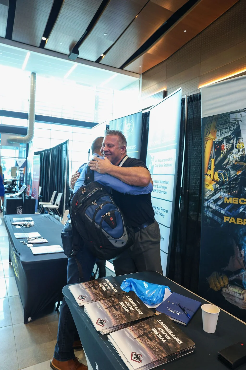 Two men hugging in a professional conference setting, with booths, banners, and conference materials visible.