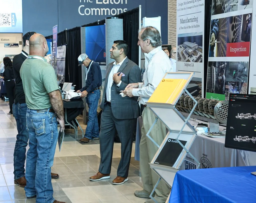 Several men are having a discussion at a trade show booth with promotional banners and displays about manufacturing and inspection technology.