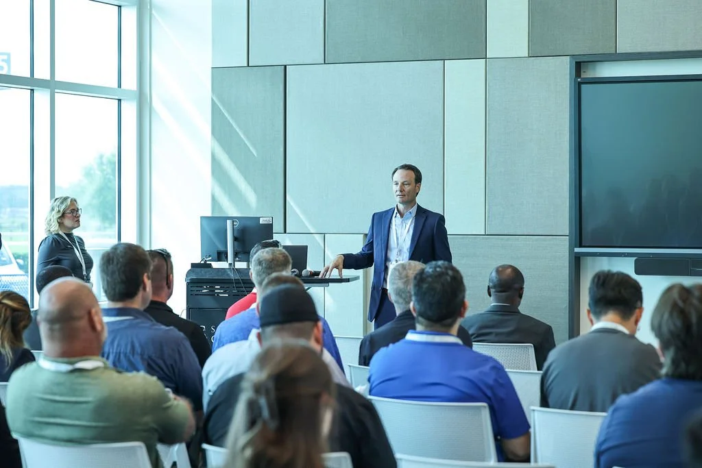 A man in a blue suit giving a presentation to a diverse group of people seated in a conference room with large windows.