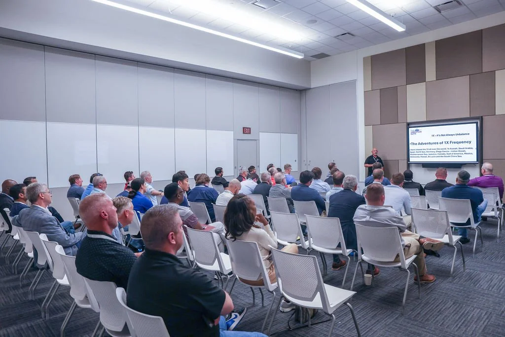 A man giving a presentation to a seated audience in a conference room, with a screen displaying a slide titled 'The Adventures of 1X Frequency'.
