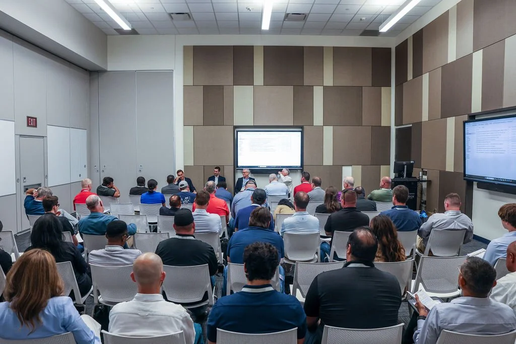 A conference room filled with attendees listening to a panel discussion at the front. There are large display screens on the wall showing presentation slides.