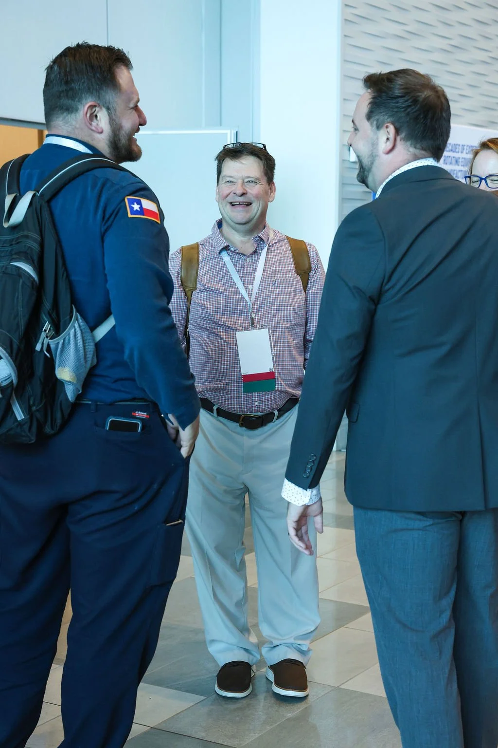 Four people engaged in conversation, three men and one woman, in an indoor setting. The woman is partially out of frame with glasses and red hair. The man in the center is smiling, wearing a checkered shirt and carrying a backpack. The man on the lef