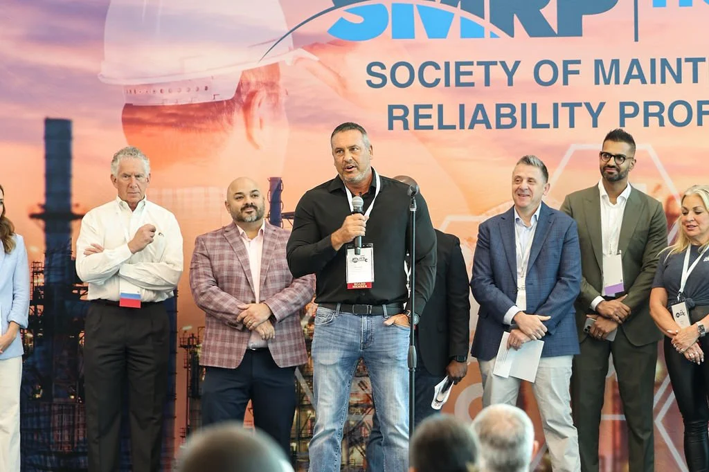Group of men and women at a conference or event on a stage, with a backdrop that reads 'SSAR' and 'Society of Maintenance Reliability Professionals.' One man is speaking into a microphone.