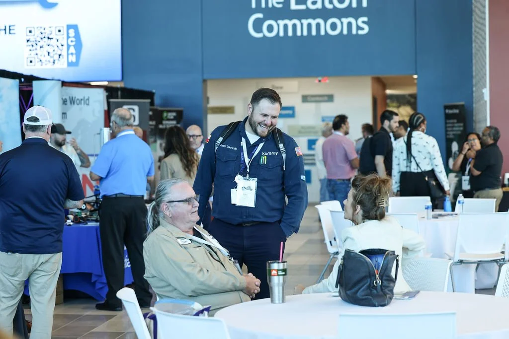 People engaging in conversation at an indoor event or conference, with a large sign reading 'The East Commons' in the background.