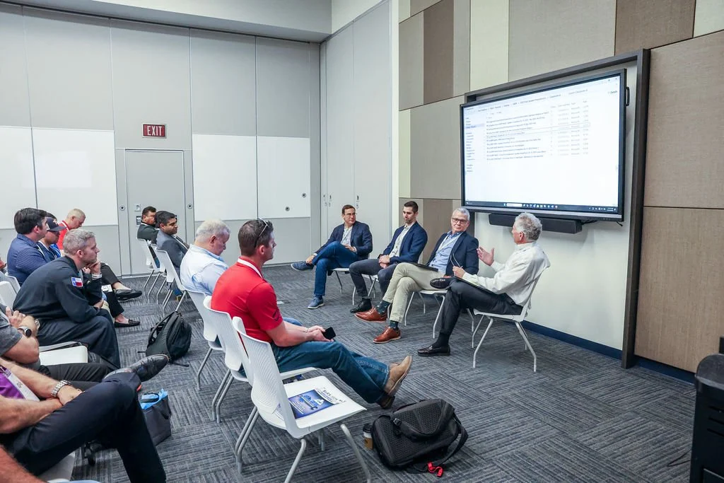 A group of people attending a panel discussion or presentation in a conference room, with five speakers sitting in front of a large screen displaying a computer screen or presentation.