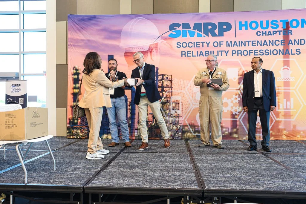 A woman in a beige suit is presenting an award to a man in a suit on a stage at a conference. Three other men are standing nearby, one holding a tablet, against a backdrop that reads 'SMRP Houston Chapter Society of Maintenance and Reliability Profes