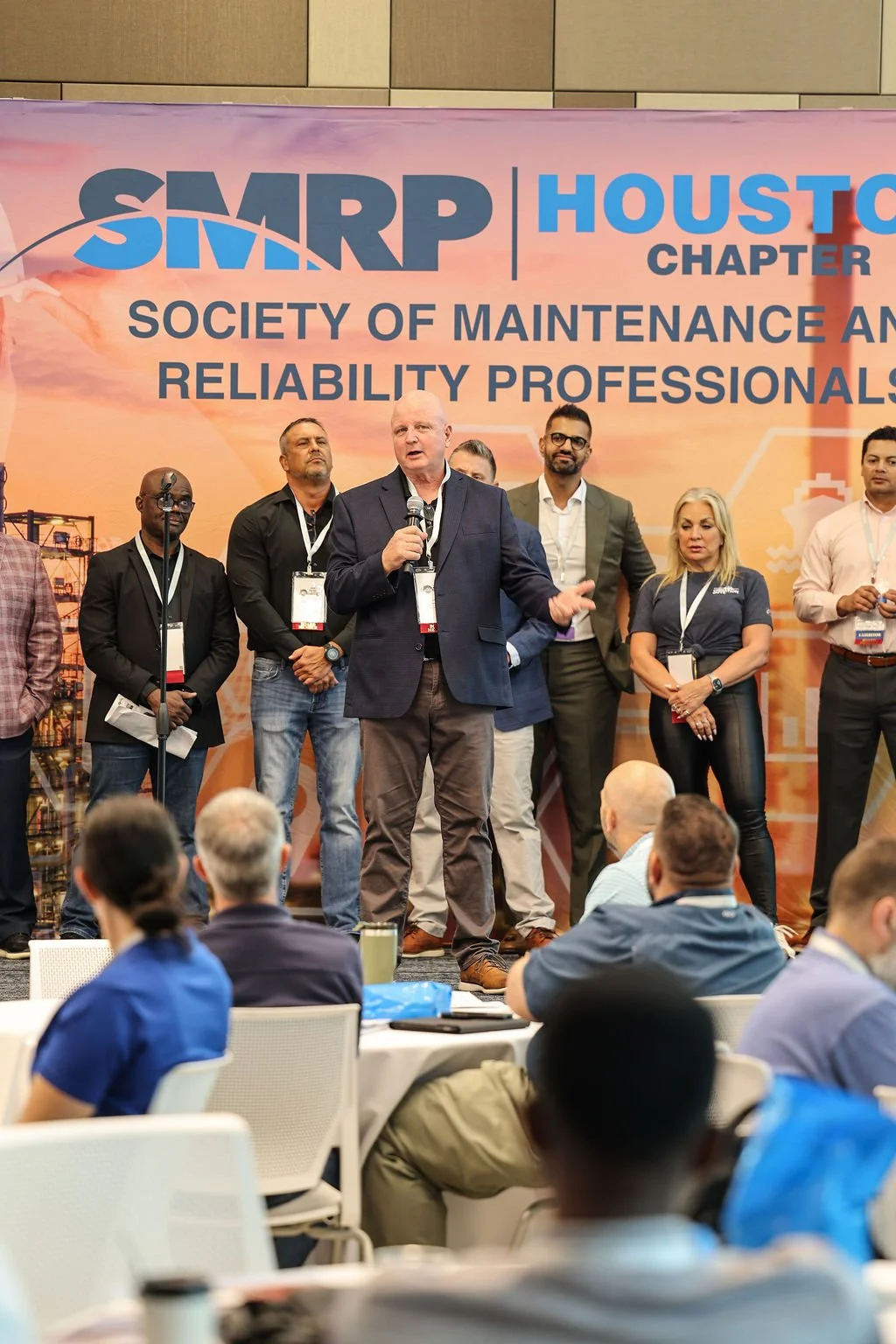 A man speaking into a microphone on stage at a conference, with the banner behind him reading "SMRP Houston Chapter Society of Maintenance and Reliability Professionals." Several people stand behind him, and attendees sit in front of the stage.