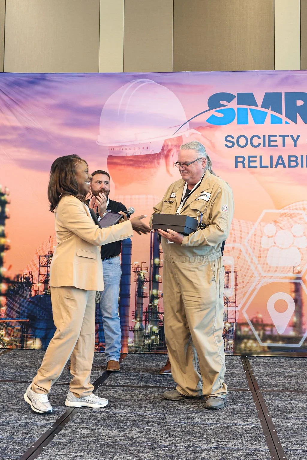 A woman in a tan suit is handing an award to a man in a tan jumpsuit on stage during a conference, with two men watching and clapping in the background.