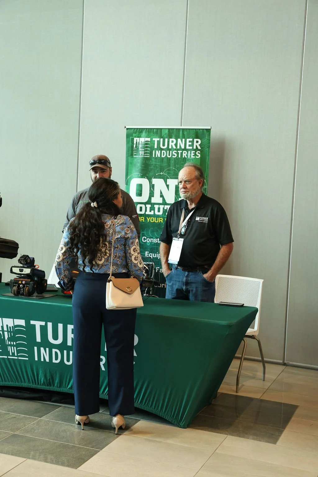 Three people at a booth with a green banner for Turner Industries, discussing. Booth has promotional materials and equipment on the table.