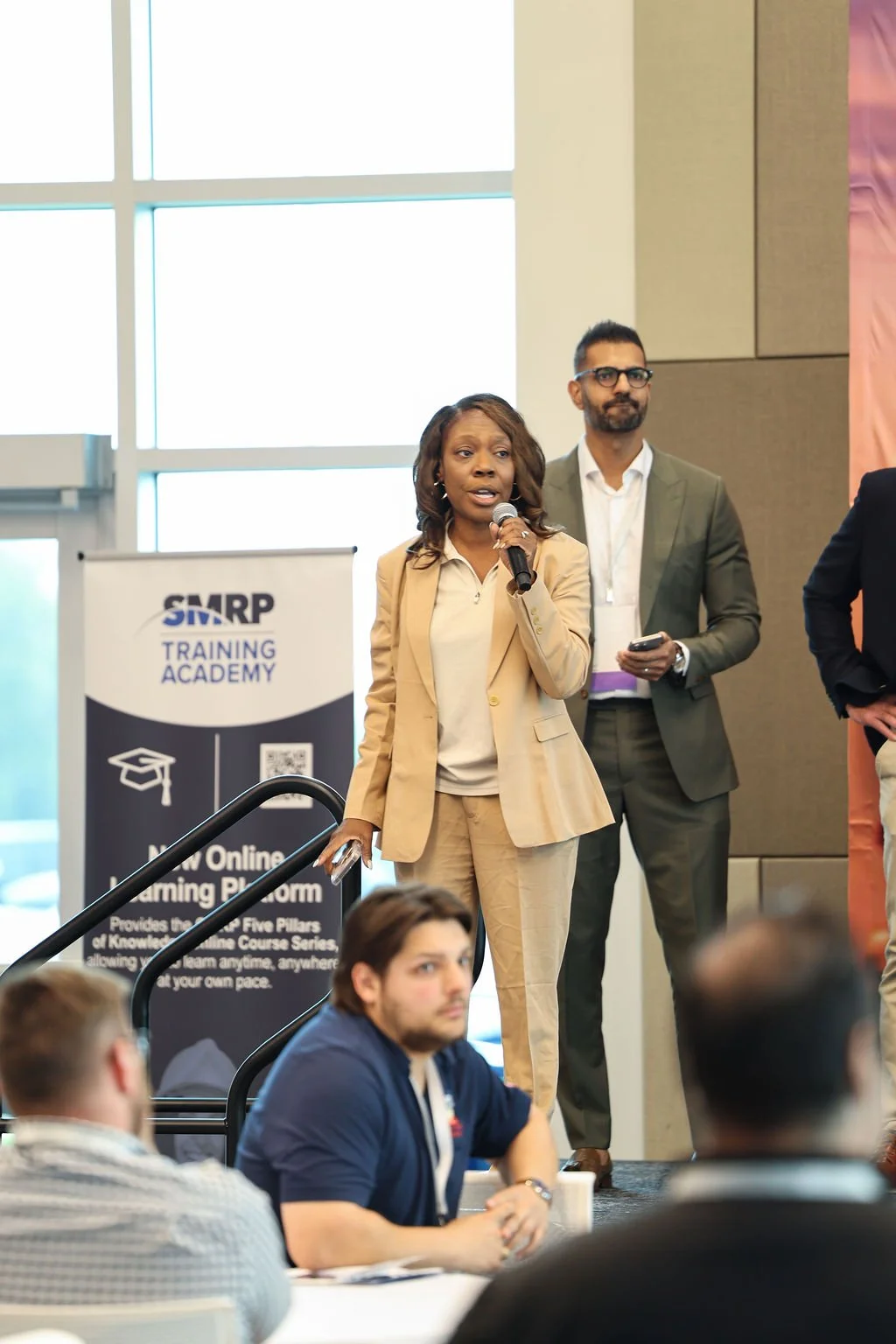 A woman speaking into a microphone at a conference, with a man standing behind her, and people seated in front. The background includes a banner for SMARP Training Academy and large windows.