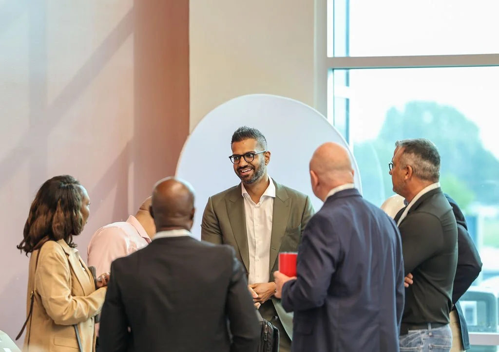 A diverse group of six professionals in a business setting, engaging in a discussion near a large window, with a man smiling at the center.