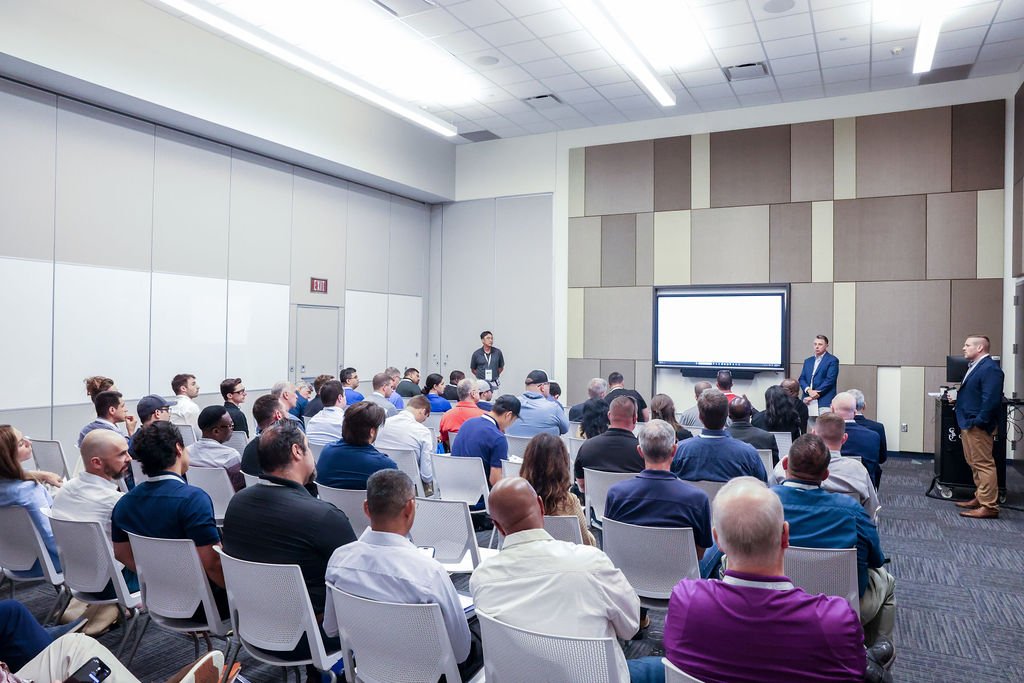 A diverse group of people attending a conference or seminar in a modern conference room, with two presenters at the front, one standing next to a large screen displaying a presentation.