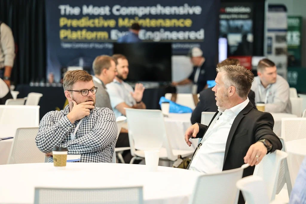 Two men seated at a conference table in conversation, with one resting his chin on his hand, and others in the background participating in a discussion, in a professional setting with a large screen and promotional banners.