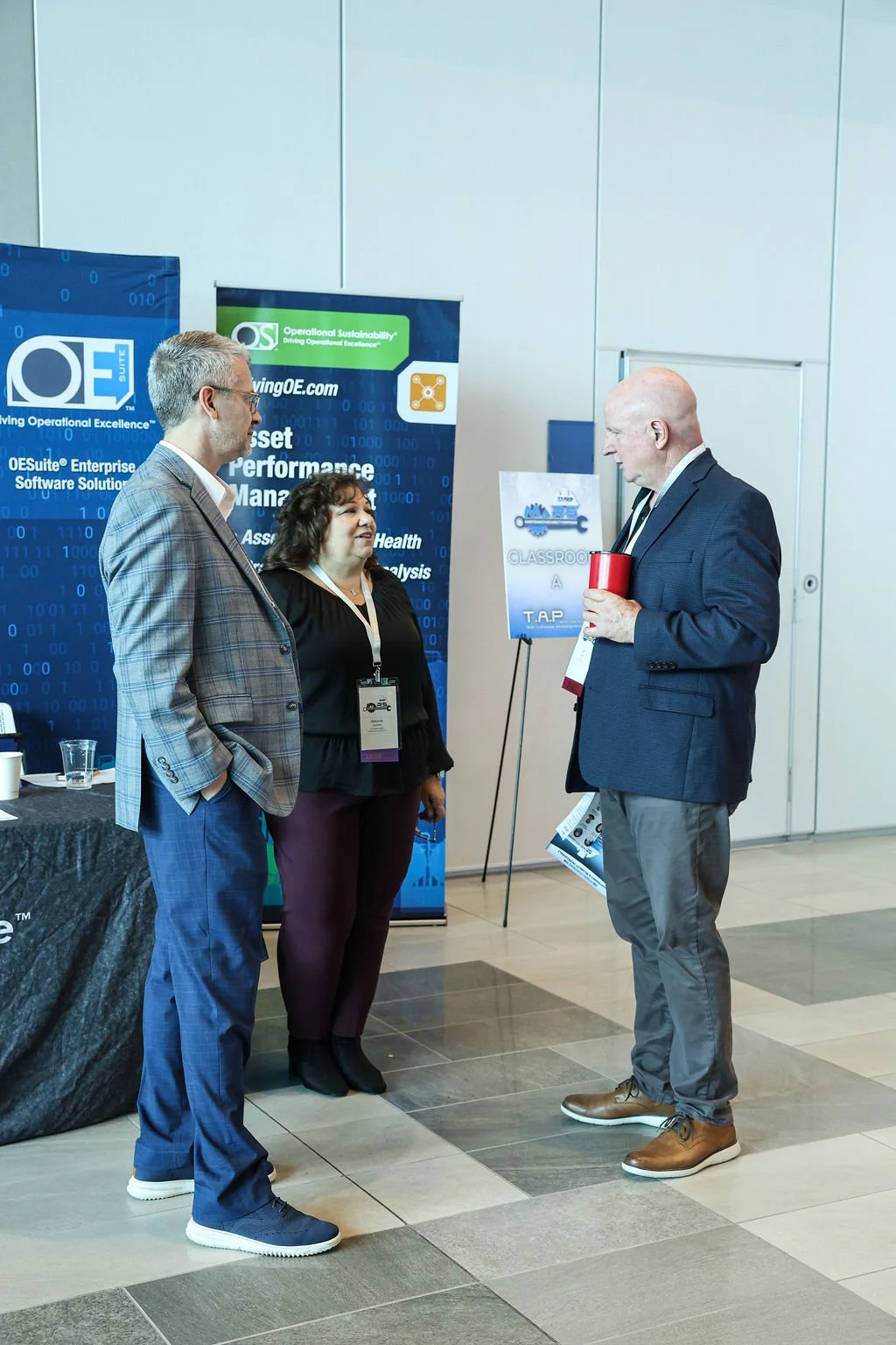 Three people talking at a conference, two men and one woman. The setting includes booth banners for QESuite Enterprise Software Solutions and Operational Sustainability.