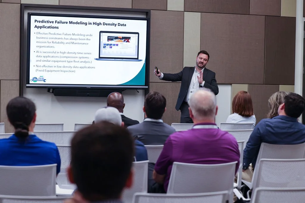 A man giving a presentation on predictive failure modeling to an audience in a conference room, with a slide displaying text and an image of a computer screen projected behind him.