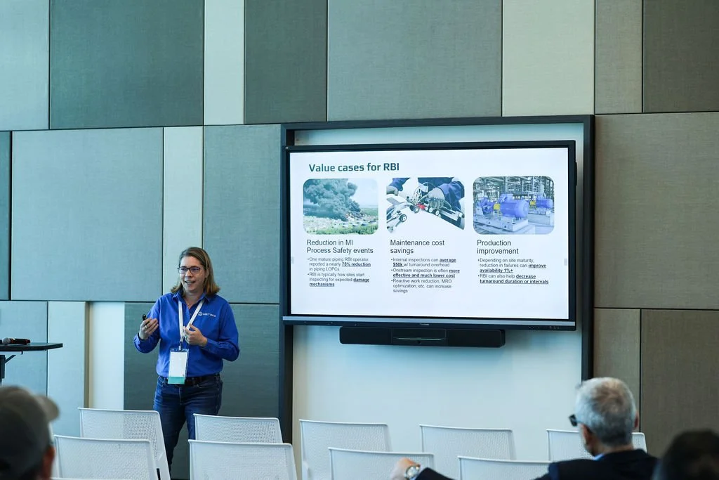 A woman giving a presentation in front of a digital screen displaying information about value cases for RBI, including reduction in MI process safety events, maintenance cost savings, and production improvement, with an audience seated and listening.