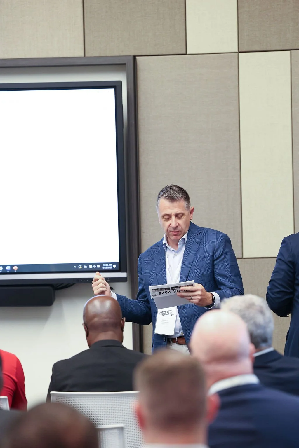 A man in a blue suit presenting in front of an audience during a conference or meeting, holding a booklet or folder, with a large blank screen behind him.