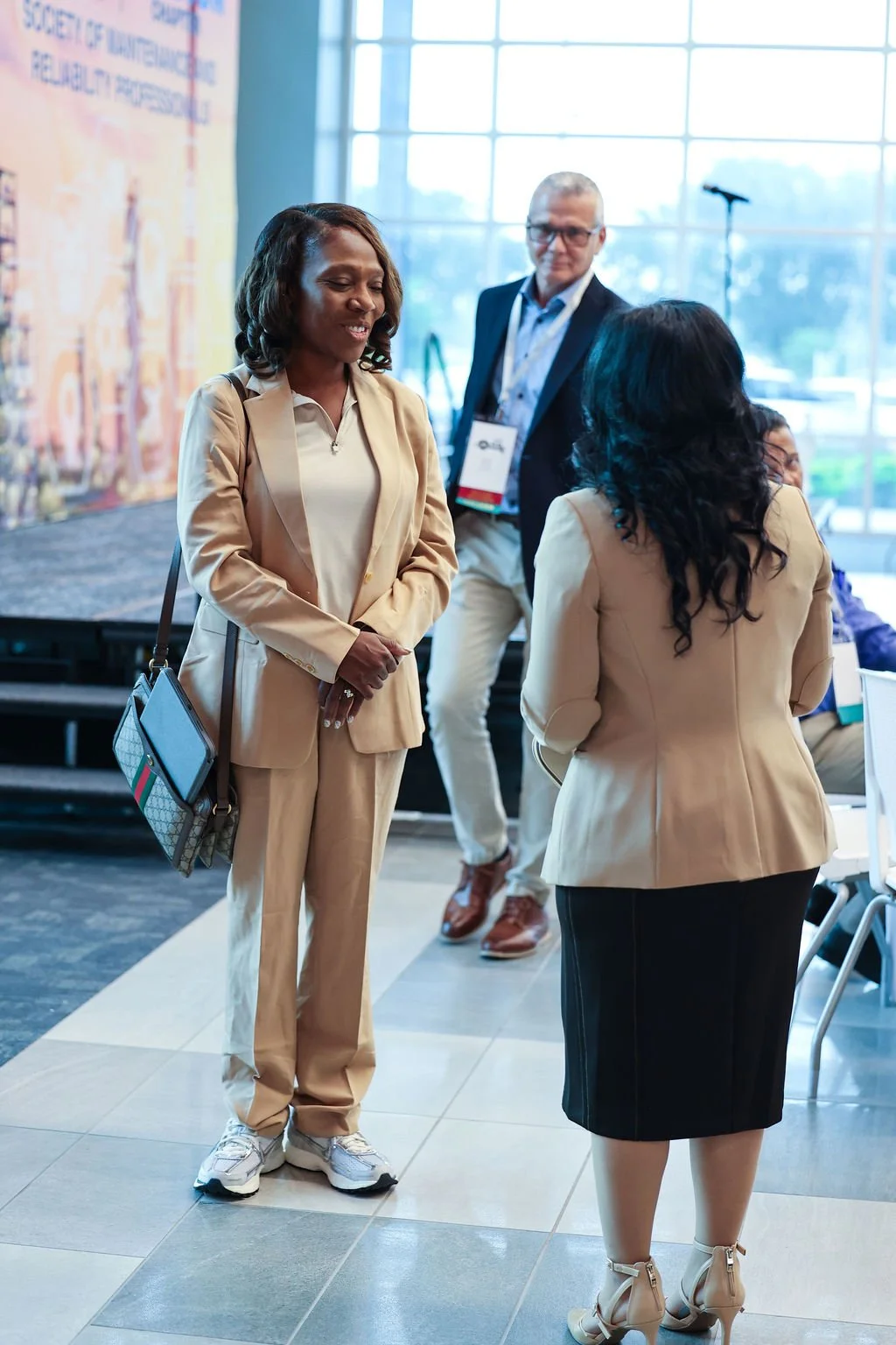 Three professionals engaging in a conversation at a conference or networking event, with large windows and a stage in the background.