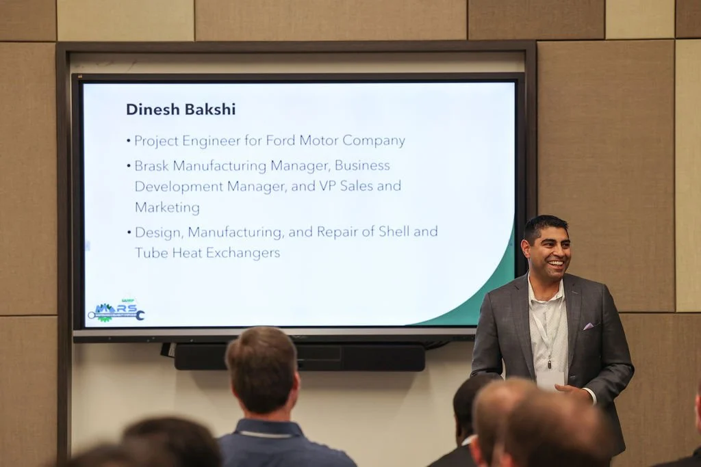 A man in a suit giving a presentation in a conference room. His slide shows his name, Dinesh Bakshi, and lists his professional roles including project engineer, manufacturing manager, and design engineer.