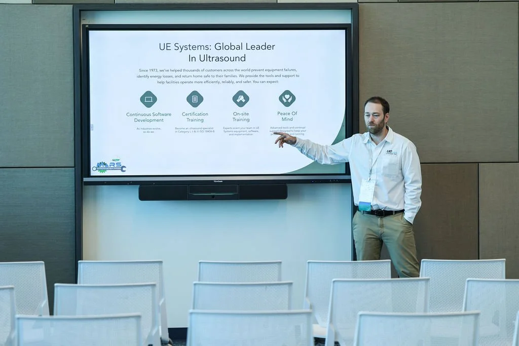 A man presenting in front of a large screen with a conference room setup, empty white chairs, and a wall behind him.