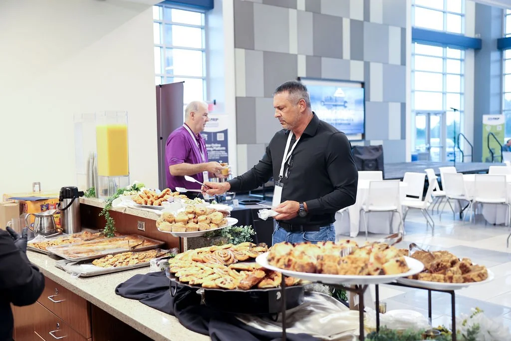 A man in a black shirt and jeans serving himself food at a buffet table with various pastries and baked goods in a modern conference center.