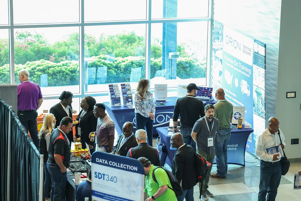 Conference or trade show with multiple booths and attendees inside a modern building with large windows and outdoor greenery.