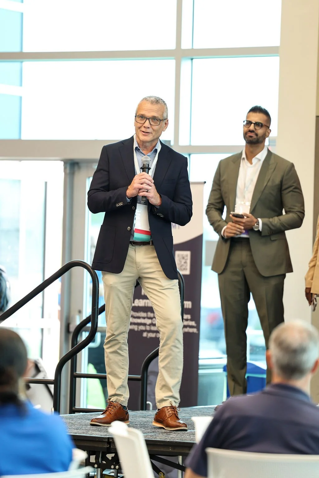 A man with glasses speaking into a microphone on stage at a conference, wearing a navy blazer, light collared shirt, beige pants, and brown shoes, with another man in a gray suit and glasses standing beside him holding a phone.