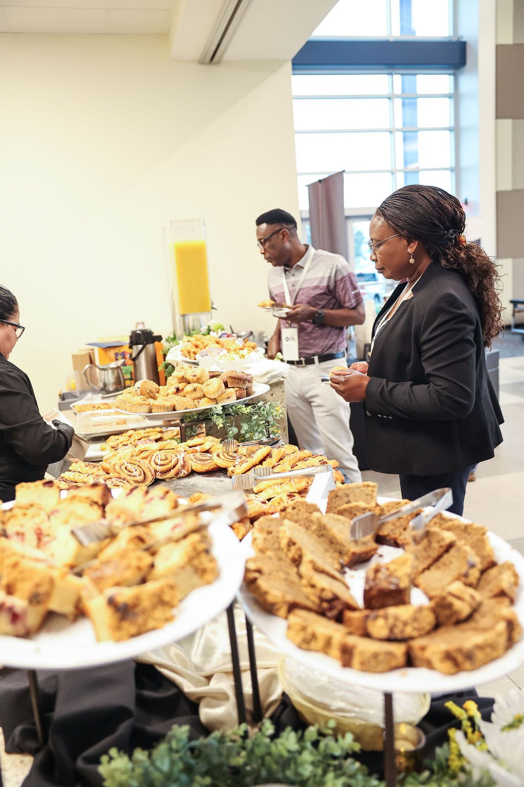 People at a buffet table with a variety of baked goods, pastries, and a tall glass of orange juice in a bright, modern room.