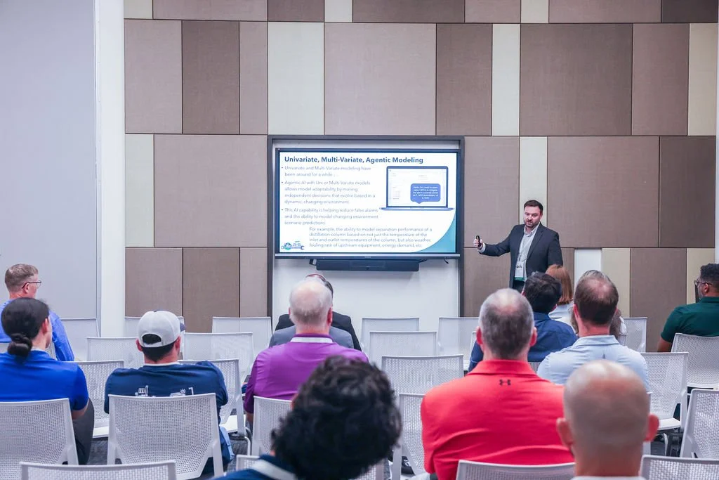 A man in a suit giving a presentation in front of an audience in a conference room with a large screen displaying a PowerPoint slide titled 'Univariate, Multi-Variance, Agentic Modeling'. The audience is sitting in white chairs, listening attentively