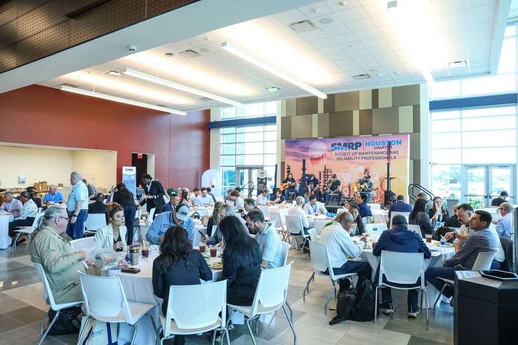 Large indoor conference with round tables, people eating and conversing, a stage at the back with a band, and a banner that reads 'SMRP Houston Chapter Society of Maintenance and Reliability Professionals'.