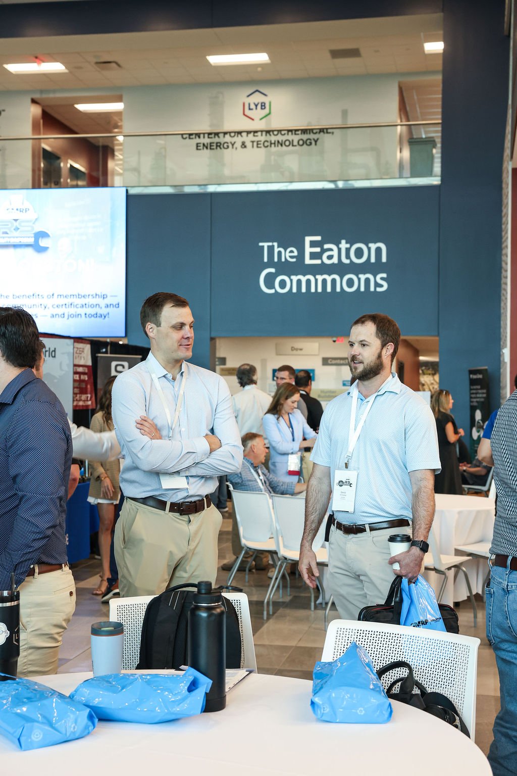 At a conference, two men are engaged in conversation in front of a sign that reads 'The Eaton Commons.' Other attendees are seated at tables and standing in the background.