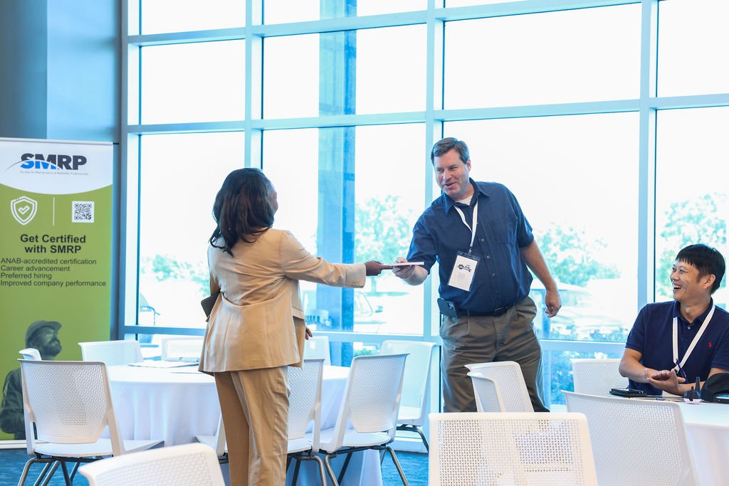A woman in a beige suit is handing a document to a man in a blue shirt during a business conference, with another man sitting at a table smiling nearby, in a bright room with large windows and a promotional banner in the background.