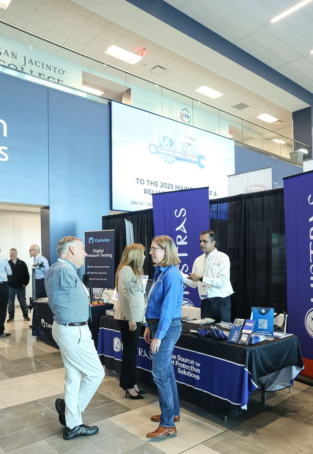 Business professionals conversing at a trade show booth with banners for SIRS and Cumulus, at an indoor conference, with a large screen displaying event information in the background.
