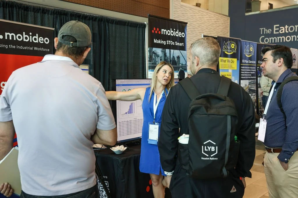 A woman in a blue dress is explaining data to three men at an industry conference booth with banners for Mobidev and other companies in the background.