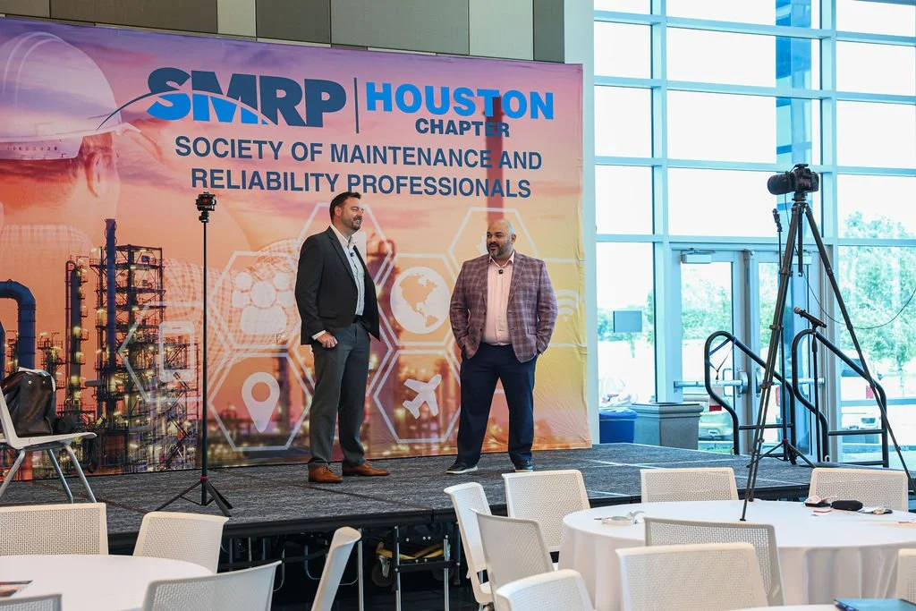 Two men are standing on a stage in front of a colorful banner at a conference. The banner reads 'SMRP Houston Chapter Society of Maintenance and Reliability Professionals'. The stage has a camera on a tripod and a microphone stand. There are empty ch