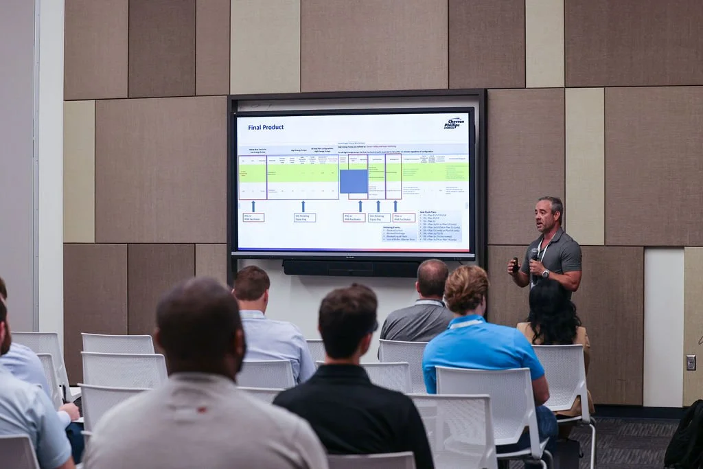A man is giving a presentation to a group of people in a conference room. He is holding a microphone and a remote control, standing next to a digital screen displaying a chart titled 'Final Product'. The audience is seated in white chairs facing the 