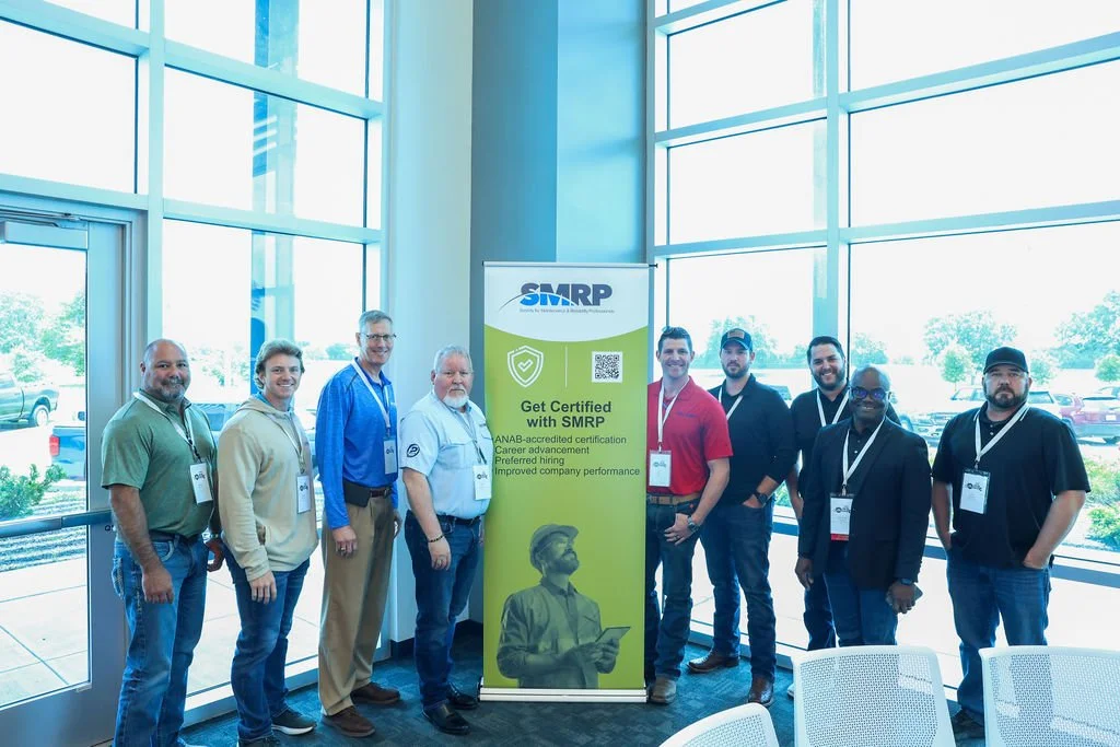 Group of nine diverse people standing indoors near a promotional banner for SMRP certification, with large windows showing a parking lot and trees outside.