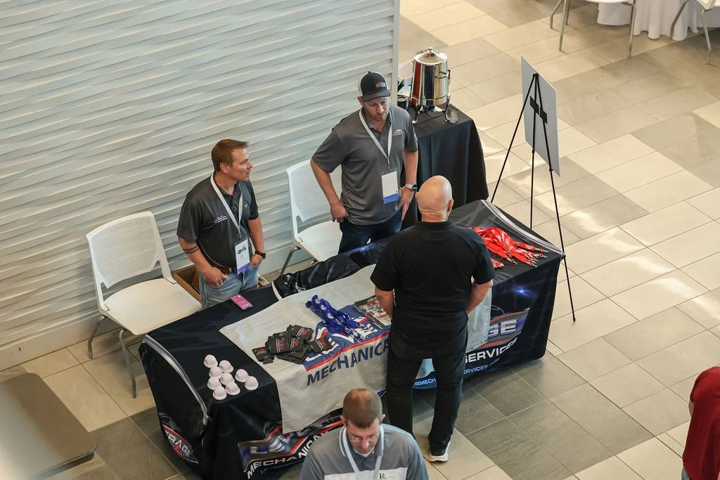 People at a booth with promotional materials, drinks, and a sign with a wrench icon at a conference or event.