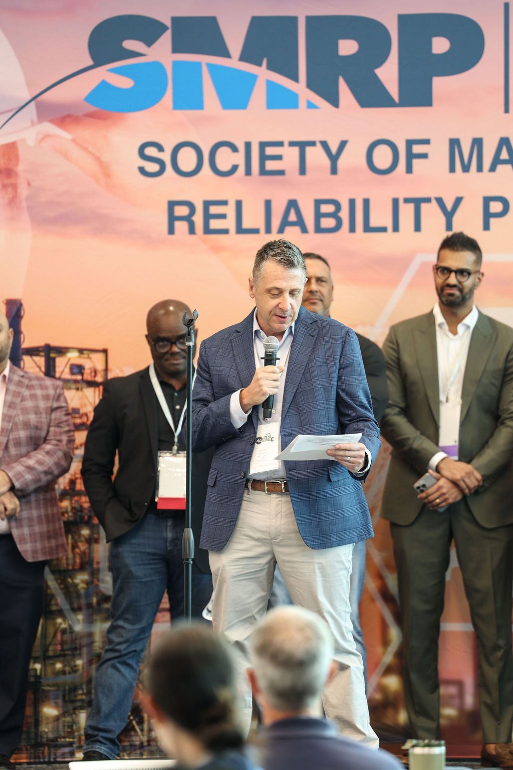 A man in a blue checked blazer speaking into a microphone at a conference with a large backdrop that reads 'SILAR' and 'Society of Materials Reliability'. Several other men are standing behind him, listening attentively.