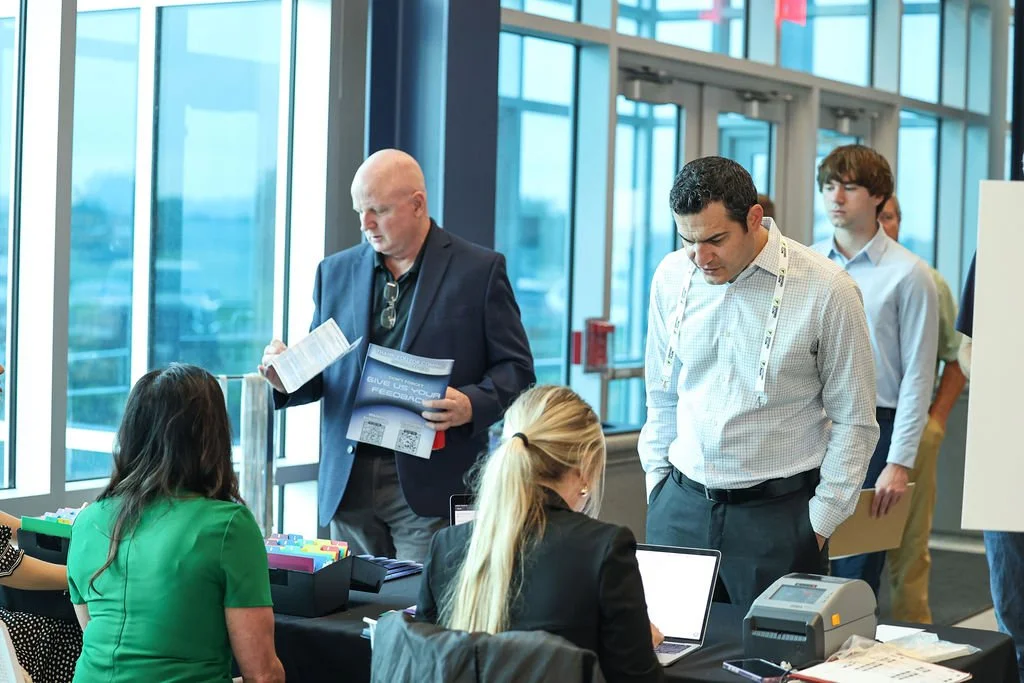 People at a registration or check-in desk inside a conference center or airport, with large windows and a view outside, some using laptops and others holding documents.