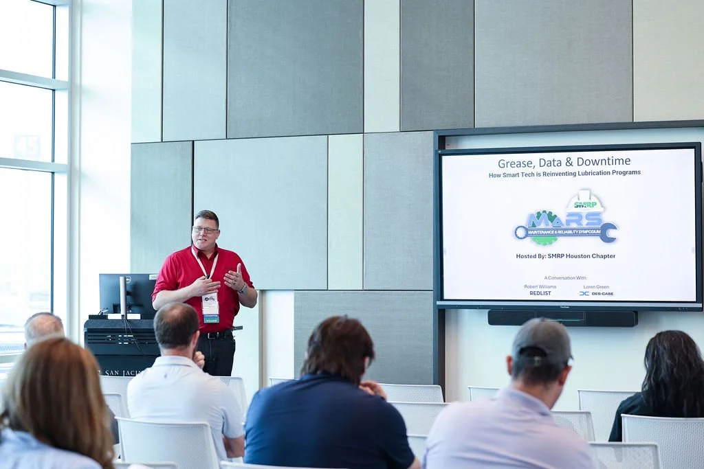 A man in a red shirt giving a presentation to an audience in a conference room with large windows. A screen displays a slide titled 'Grease, Data & Downtime' and logos for SMRP and other organizations.